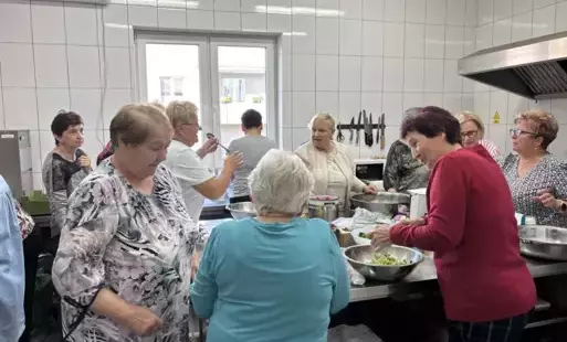 Logo Klubu Seniora. Starsza para tańcząca w ogrodzie nad nimi uśmiechnięte słońce