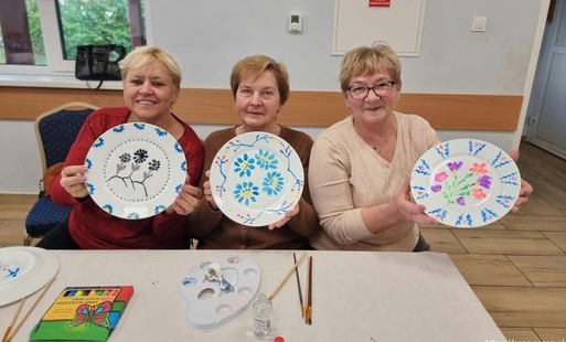 Trzy osoby siedzą przy stole podczas warsztatów plastycznych i prezentują własnoręcznie ozdobione ceramiczne talerze. Na talerzach widoczne są ręcznie malowane motywy kwiatowe w różnych kolorach i wzorach. Na stole znajdują się przybory malarskie, w tym farby, pędzle, paleta oraz inne przygotowane do dekorowania naczynia. W tle widać jasne wnętrze sali z oknem, krzesłami i ścianą z tabliczką informacyjną. Zdjęcie dokumentuje zajęcia artystyczne i efekt pracy uczestników warsztatów.